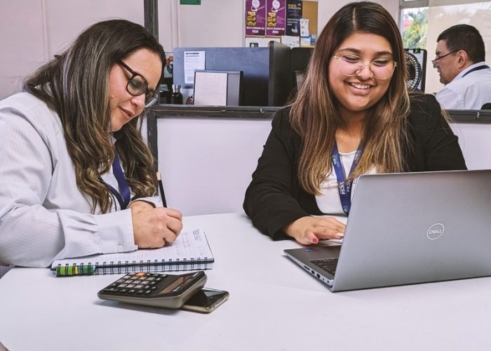 Dos mujeres conversaban en una mesa, con una computadora portátil abierta entre ellas, compartiendo ideas y colaborando.