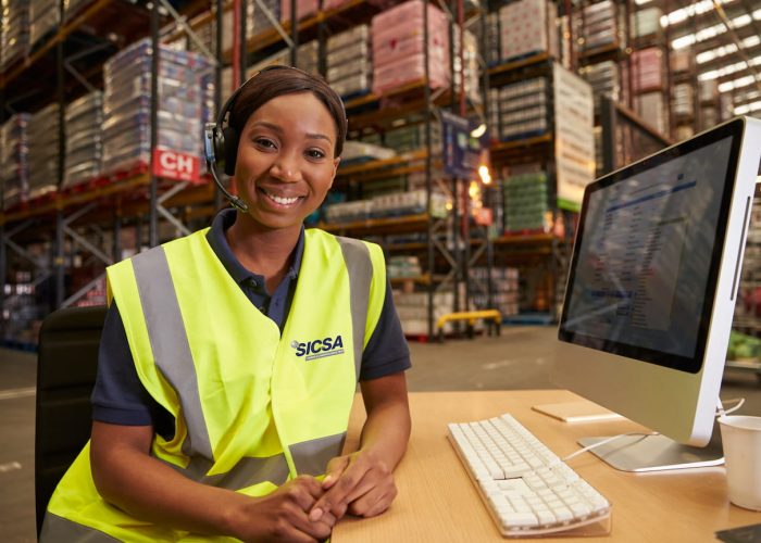 Una mujer que lleva un chaleco de seguridad está sentada en un escritorio en un almacén, concentrada en su trabajo en medio del entorno del almacén.