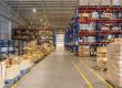 A large, organized warehouse with tall shelves holding numerous boxes and pallets, and a wide aisle running down the center.