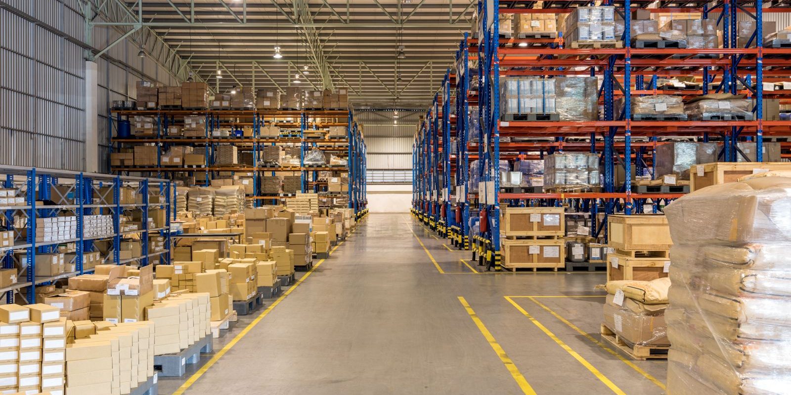 carga consolidation A large, organized warehouse with tall shelves holding numerous boxes and pallets, and a wide aisle running down the center.