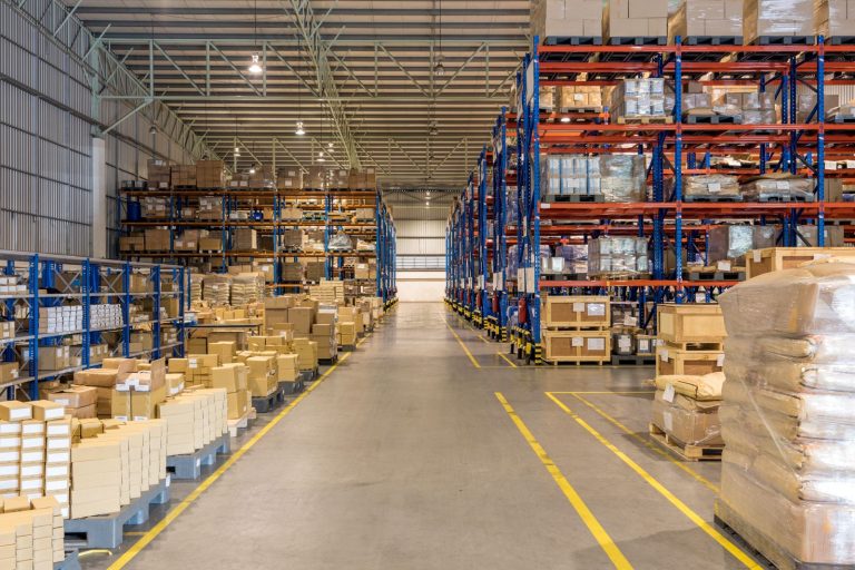 A large, organized warehouse with tall shelves holding numerous boxes and pallets, and a wide aisle running down the center.