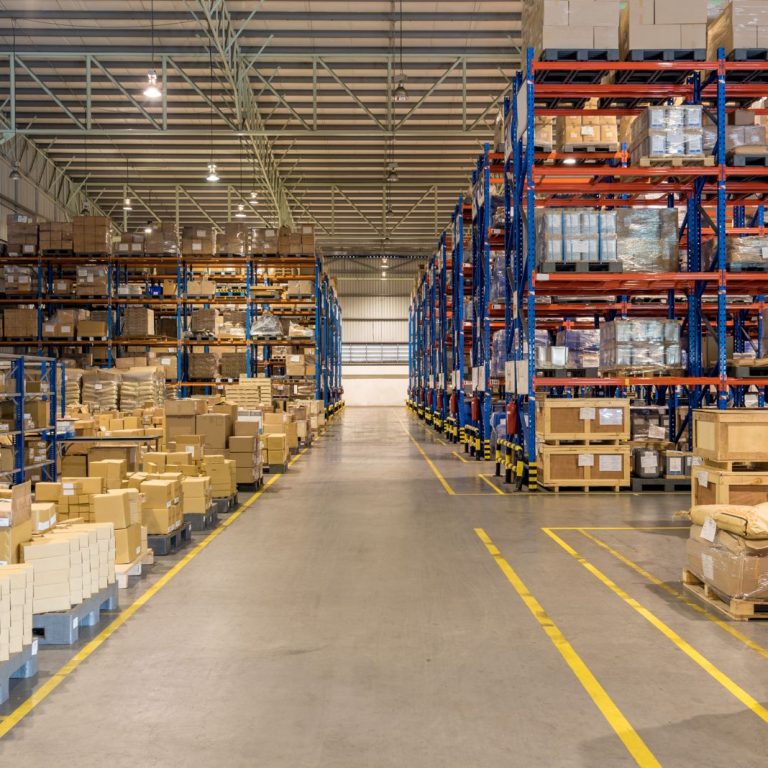 carga consolidation A large, organized warehouse with tall shelves holding numerous boxes and pallets, and a wide aisle running down the center.